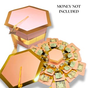 May include: Pink and gold graduation cap shaped box with a yellow tassel. The box is open and has multiple compartments for money. The text "MONEY NOT INCLUDED" is in the top right corner of the image.