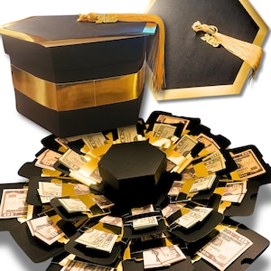 May include: A black and gold graduation-themed explosion box, with a matching graduation cap. The box is decorated with gold accents and a gold tassel. The box is filled with folded money, creating a unique gift presentation.