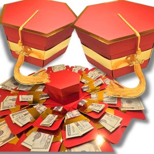May include: Two red graduation cap-shaped gift boxes with gold trim and tassels. The boxes are open, revealing a burst of red paper with cash. The gift boxes are a creative way to give money for a graduation.