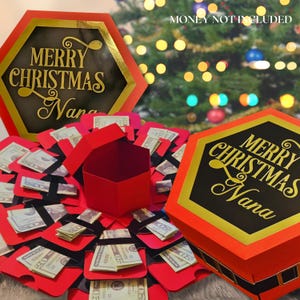 May include: A red hexagonal gift box with gold trim and the text "MERRY CHRISTMAS Nana" in gold lettering. The box is open, revealing folded money and red flaps. The words "MONEY NOT INCLUDED" are also visible.