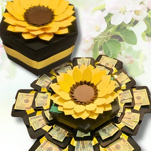 May include: A handmade papercraft gift box shaped like a sunflower. The box is black with yellow accents and a large yellow and brown sunflower on top. The box opens to reveal compartments with US twenty-dollar bills.