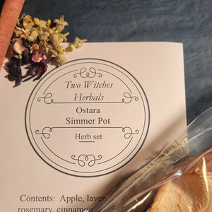 May include: A close-up of a herb set for a simmer pot. The set includes a packet of dried herbs and a card with the text "Two Witches Herbals Ostara Simmer Pot Herb set". The contents include apple, lavender, rosemary, cinnamon, and oats.