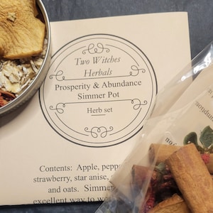 May include: A beige card with the text "Two Witches Herbals Prosperity & Abundance Simmer Pot Herb set" is next to a clear bag of herbs and a small metal bowl with dried apple slices and oats. The bag has a "Compostable" label.