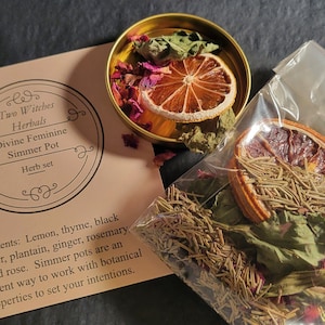 May include: A close-up of a small tin container filled with dried herbs, flowers, and citrus fruit. The tin is labeled "Two Witches Herbals Divine Feminine Simmer Pot Herb set". The text below the label reads "Contents: Lemon, thyme, black pepper, plantain, ginger, rosemary and rose. Simmer pots are an excellent way to work with botanical properties to set your intentions."