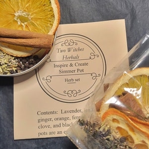 May include: A brown paper bag with the text "Two Witches Herbals Inspire & Create Simmer Pot Herb set" and a clear plastic bag containing dried orange slices, cinnamon sticks, and other herbs. The text "Compostable" is written on the plastic bag.