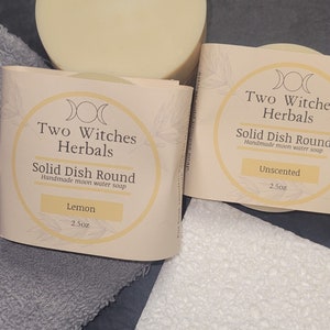 May include: Two round bars of handmade moon water soap, one labeled "Lemon" and the other "Unscented". Both bars are 2.5 ounces and feature the logo "Two Witches Herbals" with a triple moon symbol.