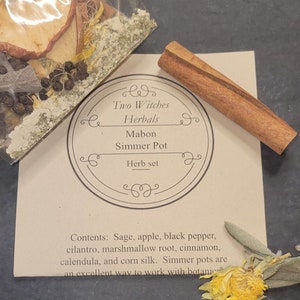 May include: A brown paper bag with a label that reads "Two Witches Herbals Mabon Simmer Pot Herb set". The bag contains dried herbs and spices, including sage, apple, black pepper, cilantro, marshmallow root, cinnamon, calendula, and corn silk. A cinnamon stick is also included.