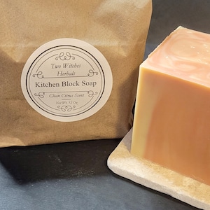 May include: A bar of kitchen block soap with a swirl of light and dark orange colors. The soap is on a light brown stone base. The soap is labeled "Two Witches Herbals Kitchen Block Soap Clean Citrus Scent Net Wt. 12 Oz."