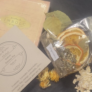 May include: A clear plastic bag with a white label that says "Compostable" contains dried orange slices, bay leaves, and herbs. The bag is sitting on a black surface next to a book with the title "Imbolc" and a small bag of oats.