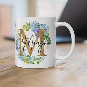 Op de afbeelding: Witte keramische mok met een gouden letter "M" en de naam "Morgan" in script, omringd door blauwe en groene bloemmotieven. De mok staat op een houten oppervlak, met een laptop op de achtergrond.