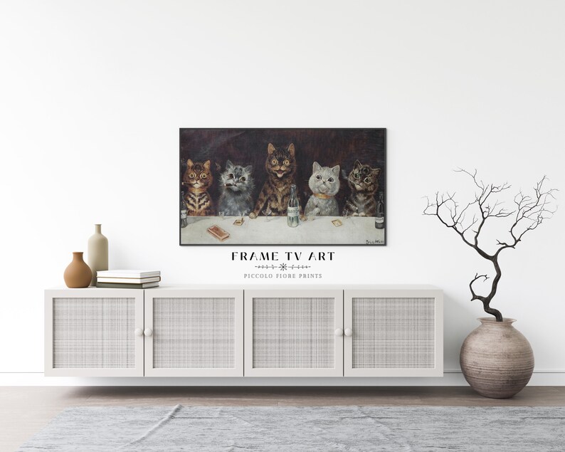 May include: A framed print of a vintage illustration featuring five cats sitting at a bar, with one cat holding a glass of beer. The print is hanging on a white wall above a white cabinet with woven rattan doors. A vase with a dried branch arrangement is on the right side of the cabinet.