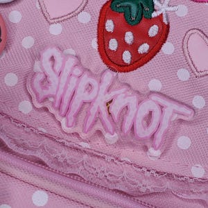 May include: Pink polka dot fabric with a pink and white embroidered strawberry and a pink plastic "Slipknot" logo.