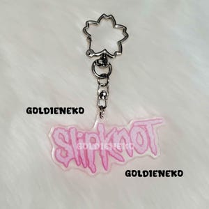 May include: A clear acrylic keychain with the word "Slipknot" in pink, outlined in a darker pink. The keychain has a silver-colored flower-shaped clasp and the text "GOLDIEKO" in black.