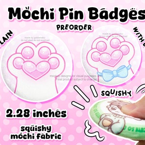 Peut inclure: Deux options de précommande pour un badge en forme de patte de chat en tissu mochi de 2,28 pouces. Une option est unie et l'autre a un nœud bleu. Le badge est fait d'un tissu mochi moelleux et est montré en train d'être pressé sur l'image.