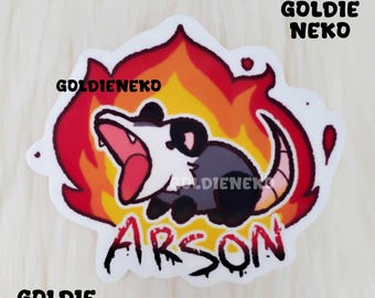 May include: A sticker featuring a cartoon possum with a red, orange, and yellow fire design. The possum is yelling and has a black eye. The text "Arson" is written in red and black below the possum. The text "Goldie Neko" is written in black at the top and bottom of the sticker.