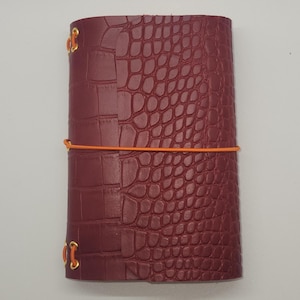 Puede incluir: Un diario de cuero burdeos con textura de piel de cocodrilo. El diario está asegurado con una banda elástica naranja y tiene detalles metálicos dorados. La cubierta es de color rojo oscuro.
