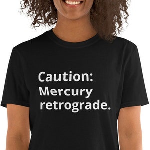 Puede incluir: Una camiseta negra con texto blanco que dice "Precaución: Mercurio retrógrado."