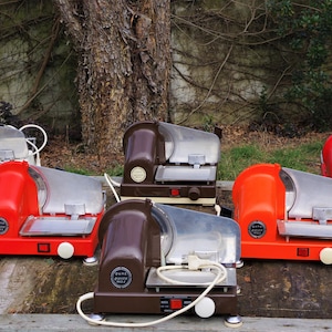 Può includere: Sei affettatrici per carne elettriche vintage di vari colori, tra cui bianco, rosso e marrone, sono allineate su una superficie di legno. Le affettatrici hanno una lama argentata e un coperchio in plastica trasparente.
