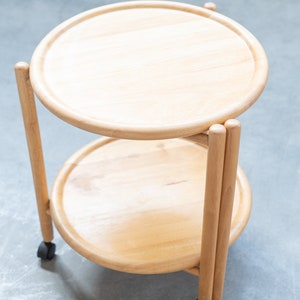 Può includere: Un carrello da servizio in legno a due livelli con ripiani rotondi e una finitura in legno naturale. Il carrello ha quattro gambe ed è su ruote.