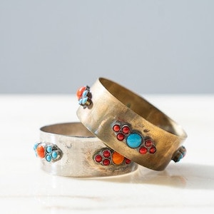 Può includere: Due braccialetti in metallo con pietre colorate. Il braccialetto superiore è in oro con pietre turchesi e rosse. Il braccialetto inferiore è in argento con pietre turchesi e rosse.