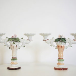 Può includere: Un paio di portacandele in ceramica bianca, ognuno con tre bocchette per candele. I portacandele sono decorati con foglie verdi, fiori rosa e dettagli marroni e dorati.