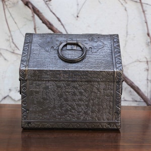 Può includere: Una piccola scatola di metallo grigio scuro con un coperchio incernierato e una maniglia ad anello di metallo decorativo. La scatola ha una superficie testurizzata ed è decorata con un motivo geometrico attorno ai bordi.