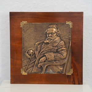 Può includere: Una placca in bronzo che raffigura un uomo barbuto seduto su una sedia. La placca è incorniciata in una cornice di legno scuro con accenti in ottone.