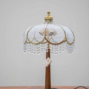Può includere: Una lampada da tavolo in cristallo e oro con un design unico e decorato. Il paralume è fatto di cristalli trasparenti e ha un bordo dorato. La base è in legno e ha una finitura nera.