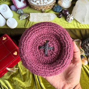 May include: A circular, deep maroon crochet object with a textured, ribbed surface and a dark gray cross-shaped stitch pattern in the center. The item is round, thick, and features a consistent yarn-based construction throughout its entire form.