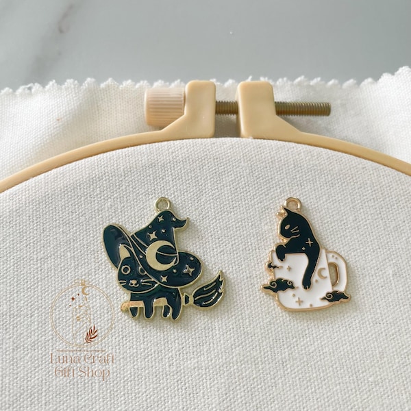 Op de afbeelding: Twee goudkleurige metalen bedels met zwart-witte emaille vulling. De ene bedel toont een zwarte kat met een heksenhoed en een maansikkel, de andere een zwarte kat in een witte mok. Beide zijn voorzien van kleine ster- en wolkdetails.