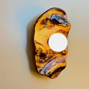 Puede incluir: Un aplique de pared con una placa trasera de forma orgánica e irregular que presenta un patrón jaspeado y arremolinado en tonos marrón oscuro, amarillo dorado y azul claro, centrado con una bombilla esférica de cristal blanco.