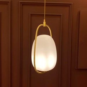 May include: A gold pendant light fixture with a white frosted glass shade hangs from a ceiling against a brown wall with paneling.