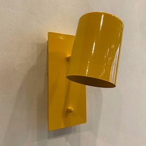 May include: A yellow wall-mounted light fixture with a cylindrical shade. The fixture is made of metal and has a simple, modern design.