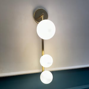 May include: A modern wall sconce with three spherical, frosted glass shades. The shades are arranged vertically along a slender, gold-toned metal rod. The fixture is mounted on a circular gold base, creating a sleek, contemporary design.