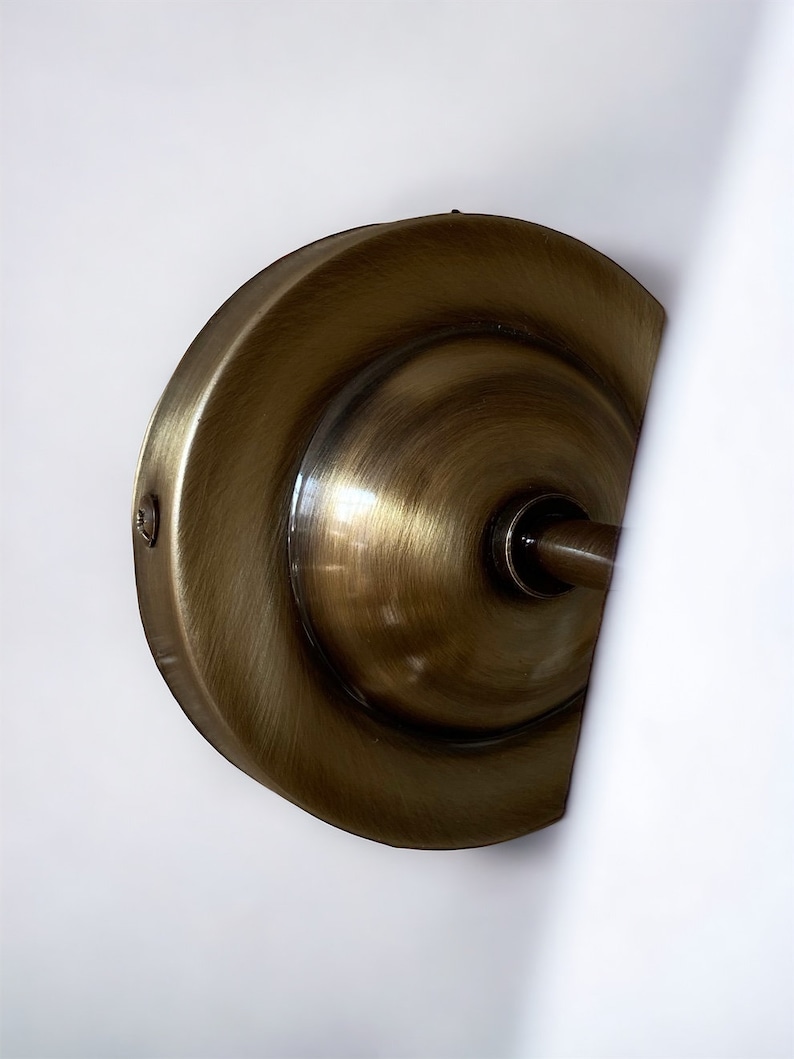 May include: A close-up of a brass wall sconce with a round, decorative backplate. The sconce has a smooth, polished finish and a slightly curved design.