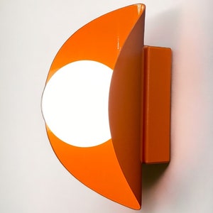 May include: Orange wall sconce with a white circular light fixture in the center. The sconce is shaped like a crescent moon.