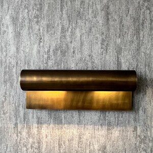 May include: A modern wall sconce with a cylindrical top and a rectangular base, both in a warm brass finish. The sconce is mounted on a textured gray wall, and emits a soft, inviting light.
