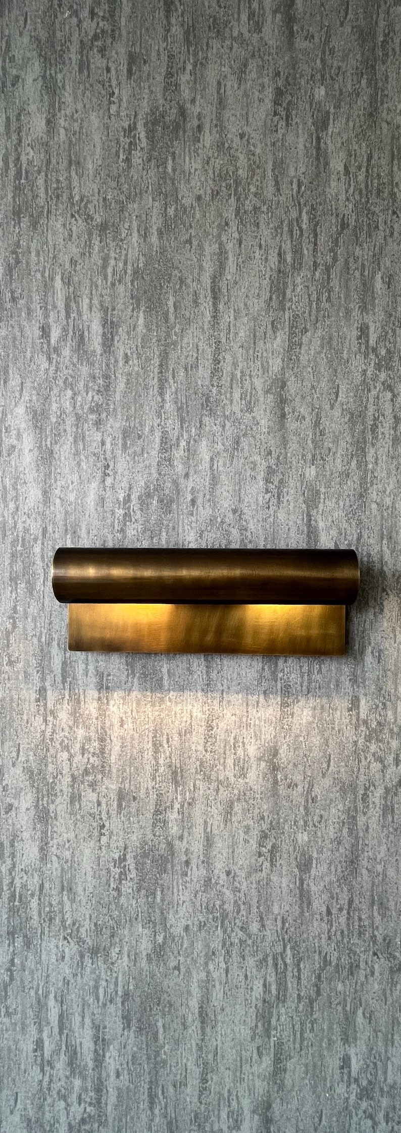 May include: A modern wall sconce with a cylindrical top and a rectangular base, both in a warm brass finish. The sconce is mounted on a textured gray wall, and emits a soft, inviting light.