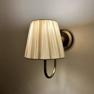 May include: A wall-mounted light fitting with a cream-coloured pleated fabric shade and a gold-toned metal base.