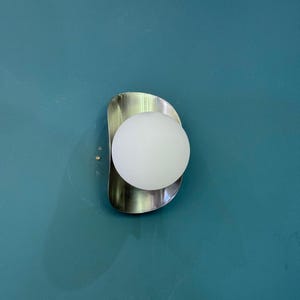 May include: A contemporary wall sconce featuring a brushed metal backplate and a spherical, frosted glass light. The fixture is mounted on a teal wall, creating a minimalist design. The light emits a soft white glow.