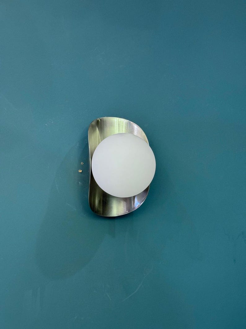 May include: A contemporary wall sconce featuring a brushed metal backplate and a spherical, frosted glass light. The fixture is mounted on a teal wall, creating a minimalist design. The light emits a soft white glow.