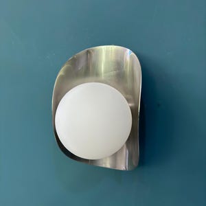 May include: A contemporary wall light with a brushed metal backplate and a spherical, frosted glass bulb. The light is mounted on a teal wall, creating a minimalist design.
