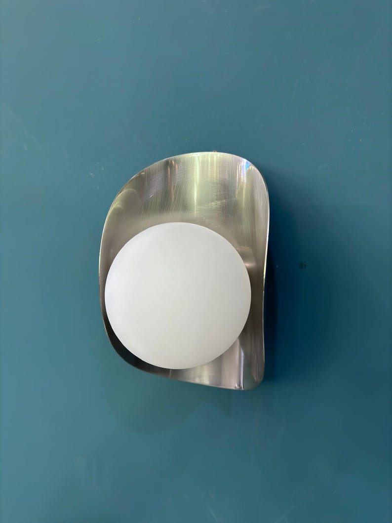 May include: A contemporary wall light with a brushed metal backplate and a spherical, frosted glass bulb. The light is mounted on a teal wall, creating a minimalist design.