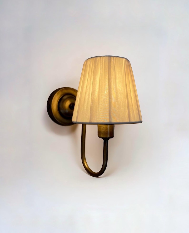 May include: A brass wall sconce with a cream-coloured fabric lampshade. The lampshade has vertical pleats and a curved bottom.