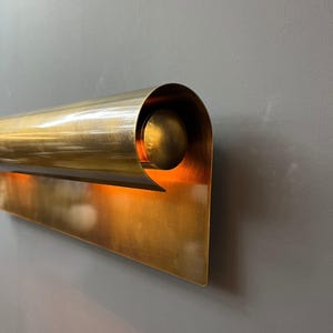 May include: A brass wall sconce with a curved, cylindrical top and a flat, rectangular base. The sconce has a warm, golden hue and is mounted on a gray wall. The design is modern and minimalist.