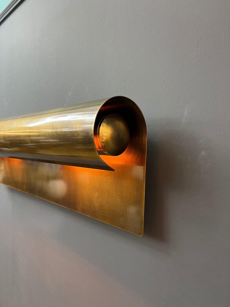 May include: A brass wall sconce with a curved, cylindrical top and a flat, rectangular base. The sconce has a warm, golden hue and is mounted on a gray wall. The design is modern and minimalist.