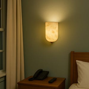 May include: A wall-mounted, cream-colored sconce with a rounded bottom emits a warm glow. The lamp is mounted on a muted green wall, above a wooden nightstand with a black telephone and remote control. A bed and window are also visible.