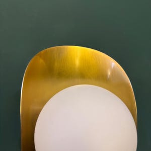 May include: A contemporary wall light with a gold-toned, curved metal backplate and a spherical, frosted white glass globe. The light is set against a dark green wall, providing a stylish contrast.