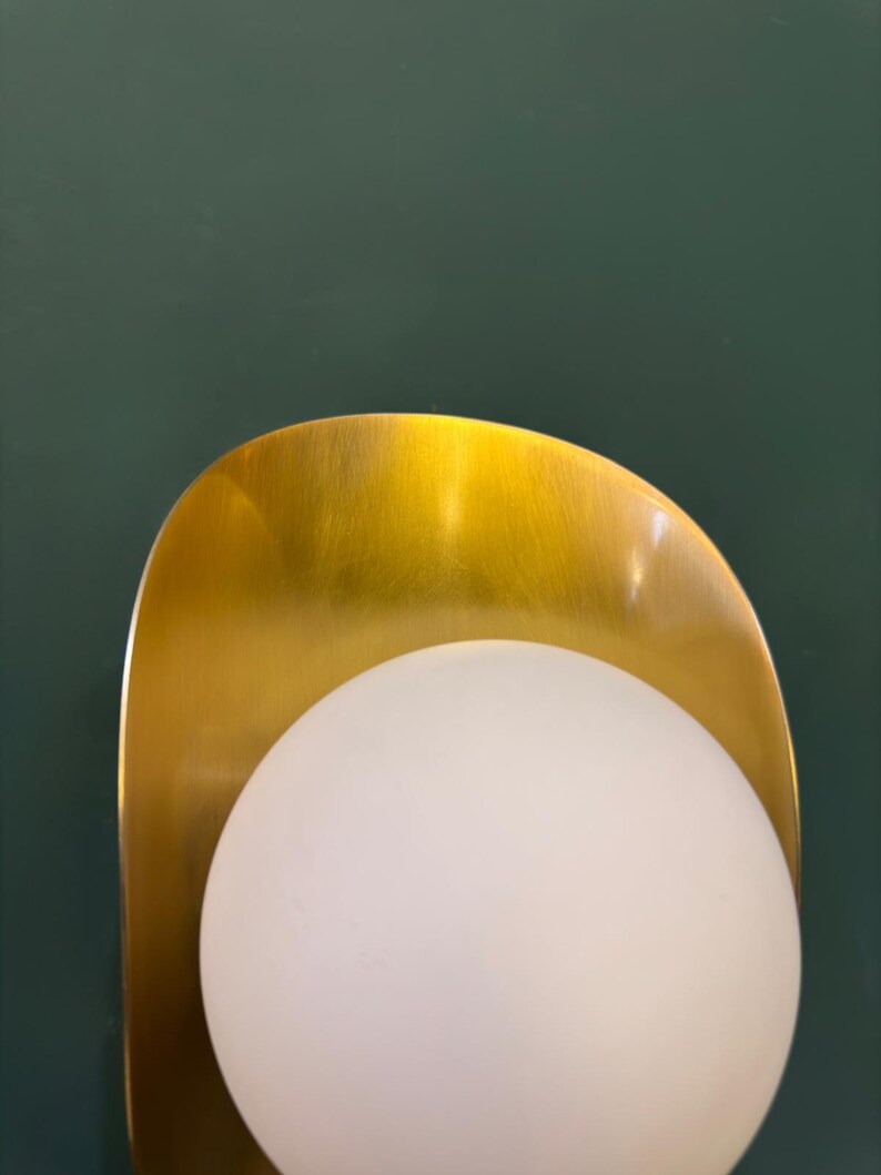 May include: A contemporary wall light with a gold-toned, curved metal backplate and a spherical, frosted white glass globe. The light is set against a dark green wall, providing a stylish contrast.