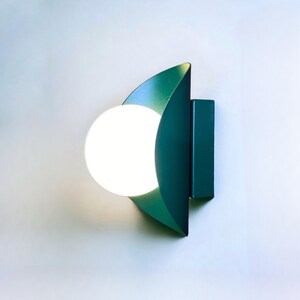 May include: A teal green wall sconce with a white globe light fixture. The sconce is shaped like a crescent moon.